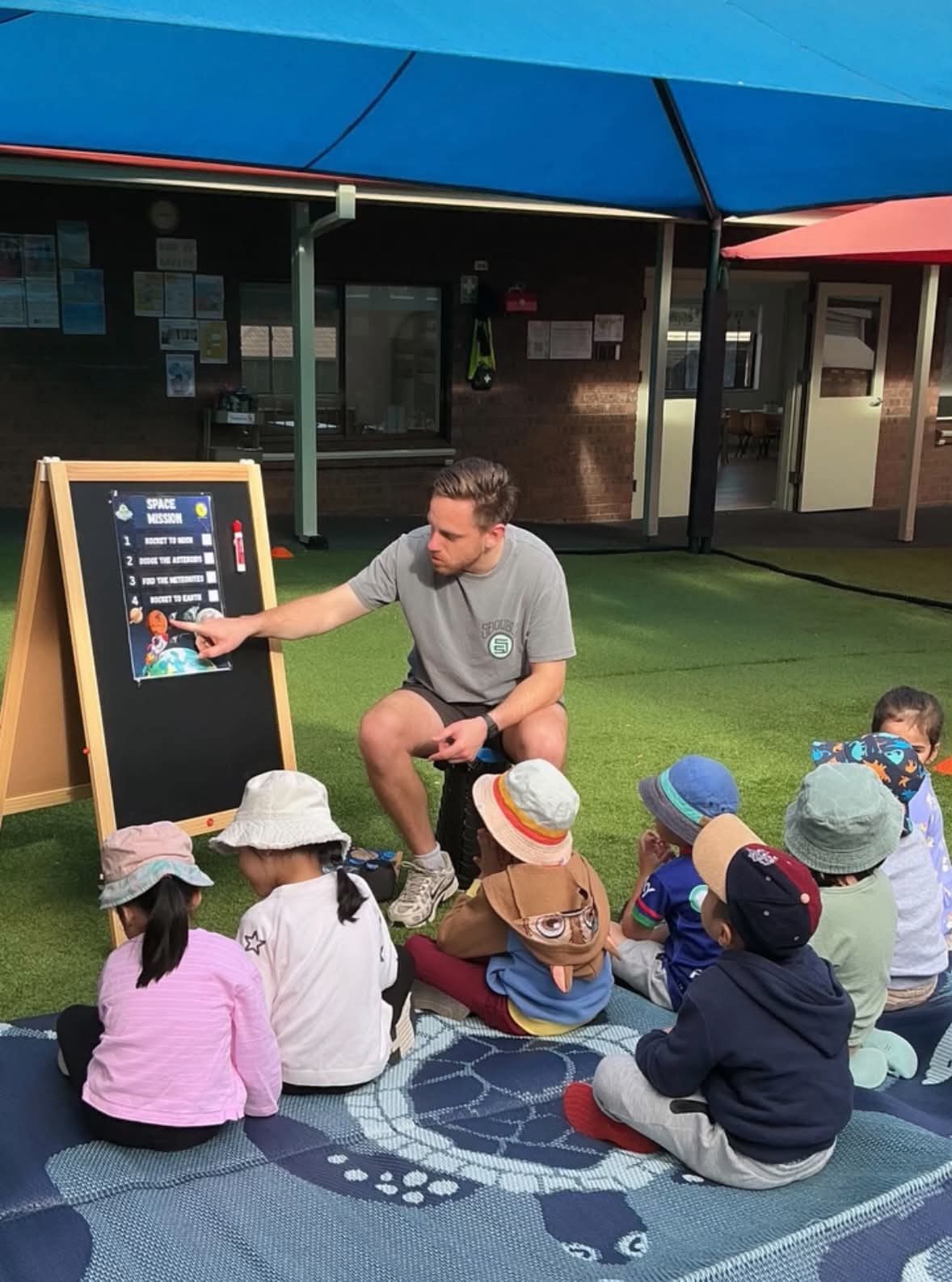 A coach pointing to the Space Mission board while preschool children sit on a mat